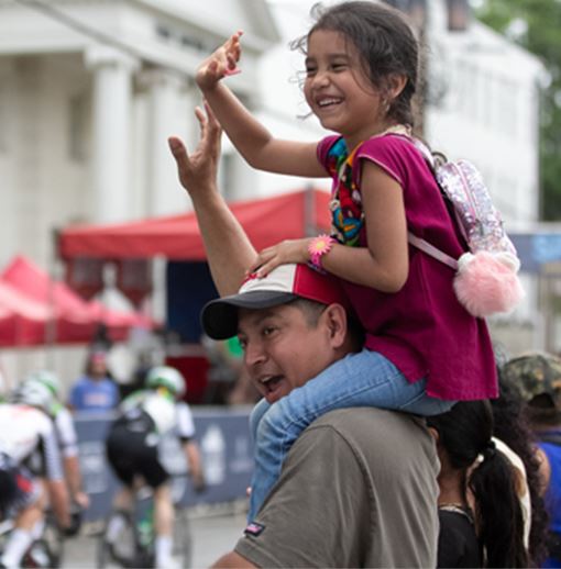 Man and his daughter who sitting on his shoulders waving at the bikers in the bike race